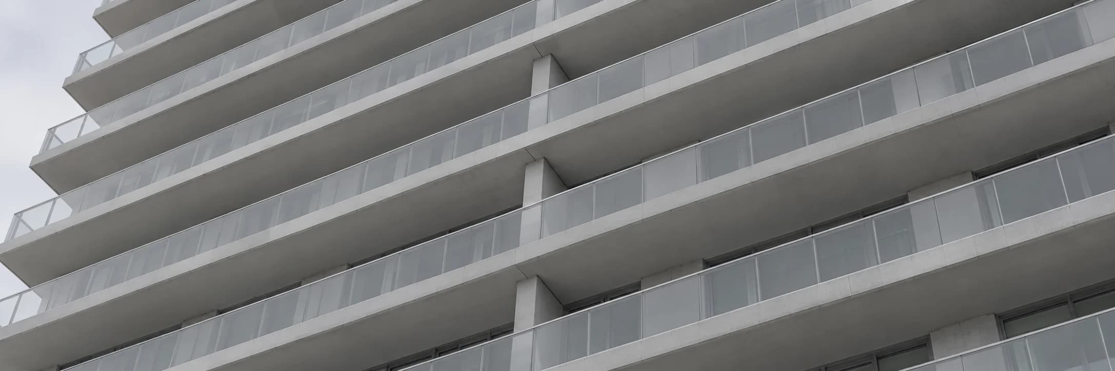 Fachada de edifício residencial vista de baixo, com varandas em fita e guarda-corpos de vidro repetidos andar a andar.